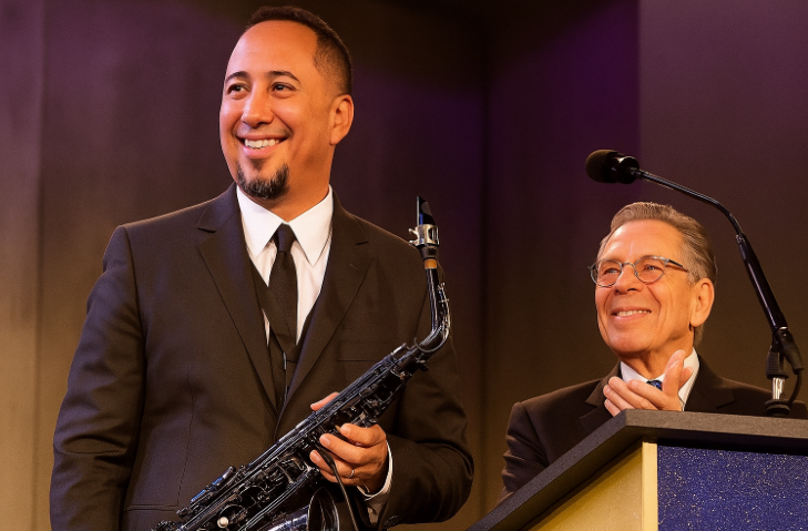 Cleto Escobedo III: Remembering Jimmy Kimmel’s Lifelong Friend and Beloved Band Leader Cleto Escobedo III smiles while holding his saxophone on stage during a live event, dressed in a black suit beside another man at a podium under warm purple lighting.